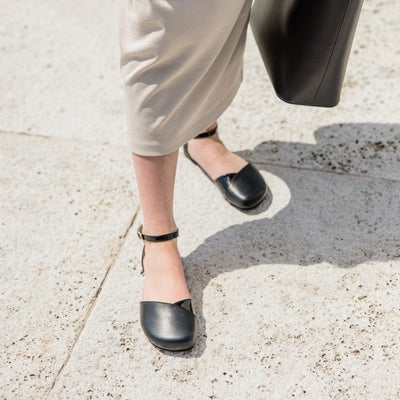 Black leather Shapen Poppy D’orsay dressy flats with a tan footbed and tan rubber soles. The flats have a heel cup held on by a dainty, patent leather ankle strap. Both shoes are shown staggered slightly above from the front on a light-skinned woman. She is standing on a stone tile pathway wearing a long beige dress holding a black leather purse on the right. #color_black