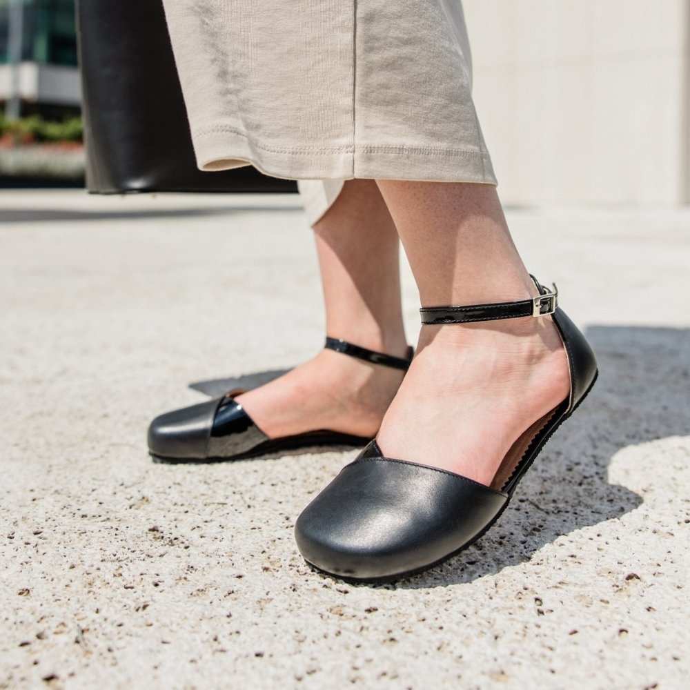 Black leather Shapen Poppy D’orsay dressy flats with a tan footbed and tan rubber soles. The flats have a heel cup held on by a dainty, patent leather ankle strap. Both shoes are shown facing diagonally left with the right foot heel popped on a light-skinned woman. She is standing on a stone tile pathway wearing a long beige dress holding a black leather purse. #color_black