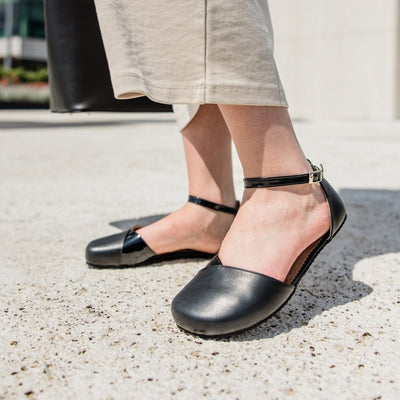 Black leather Shapen Poppy D’orsay dressy flats with a tan footbed and tan rubber soles. The flats have a heel cup held on by a dainty, patent leather ankle strap. Both shoes are shown facing diagonally left with the right foot heel popped on a light-skinned woman. She is standing on a stone tile pathway wearing a long beige dress holding a black leather purse. #color_black