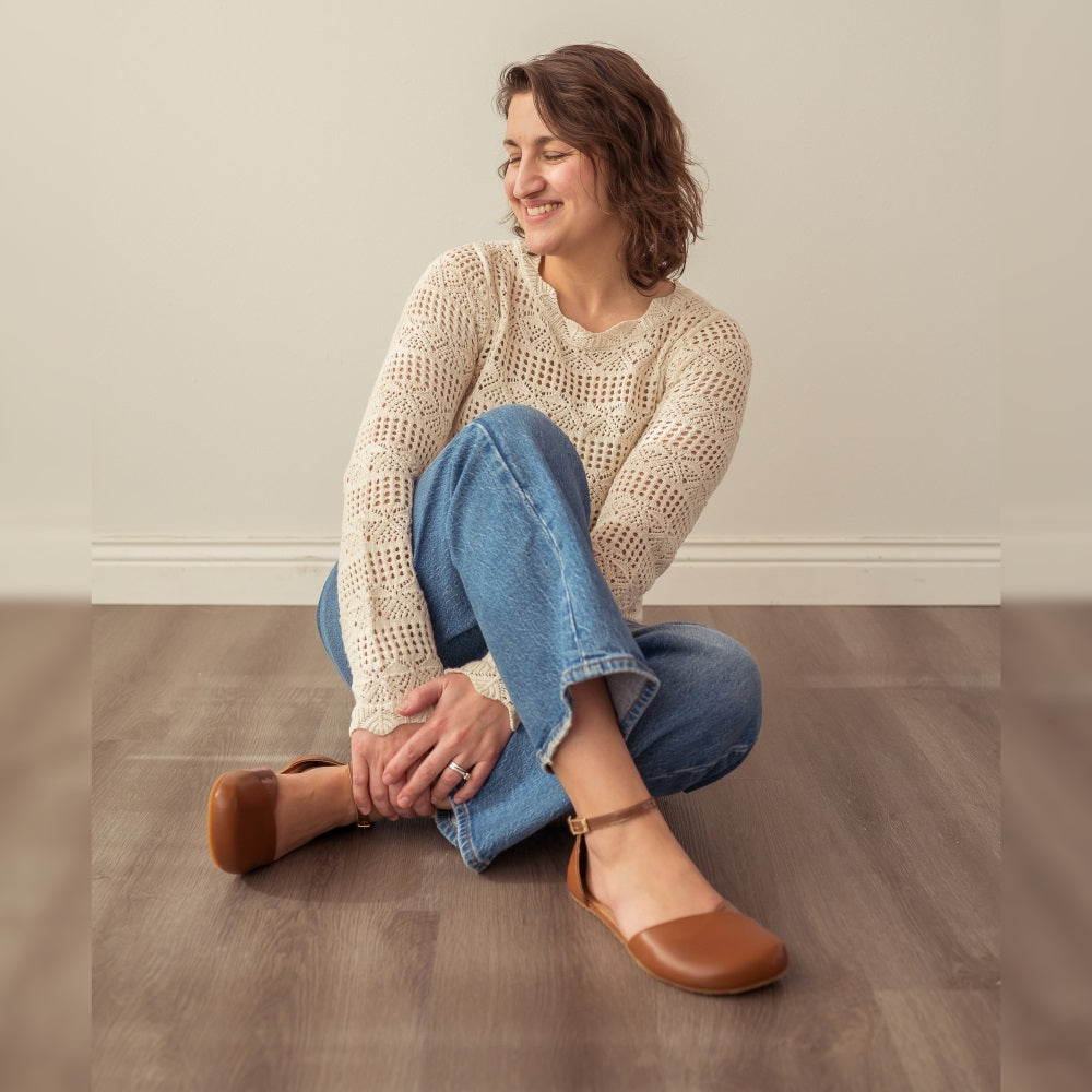A photo of Shapen Poppy D’orsay dressy flats with a leather upper and rubber soles. The flats are a cognac brown color with a heel cup and dainty ankle strap, the inside of the sole are a beige color. Both flats are shown here from the front and side on a woman wearing light-wash wide-legged jeans with an ivory crochet long-sleeve top, sitting with her right leg crossed over the left, on a grey wooden floor with a white wall in the background. #color_cognac