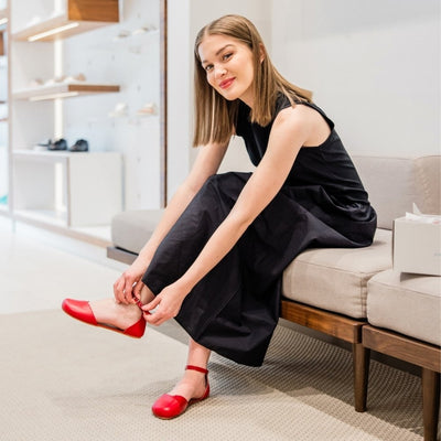 Red leather Shapen Poppy D’orsay dressy flats with a tan footbed and tan rubber soles. The flats have a heel cup held on by a dainty, patent leather ankle strap. Both shoes are shown facing left on a light-skinned woman with short, light-brown hair. She is sitting on a beige fabric and wooden bench wearing a long black dress with her left leg crossed over the right and both hands on the left shoe ankle strap. #color_cherry