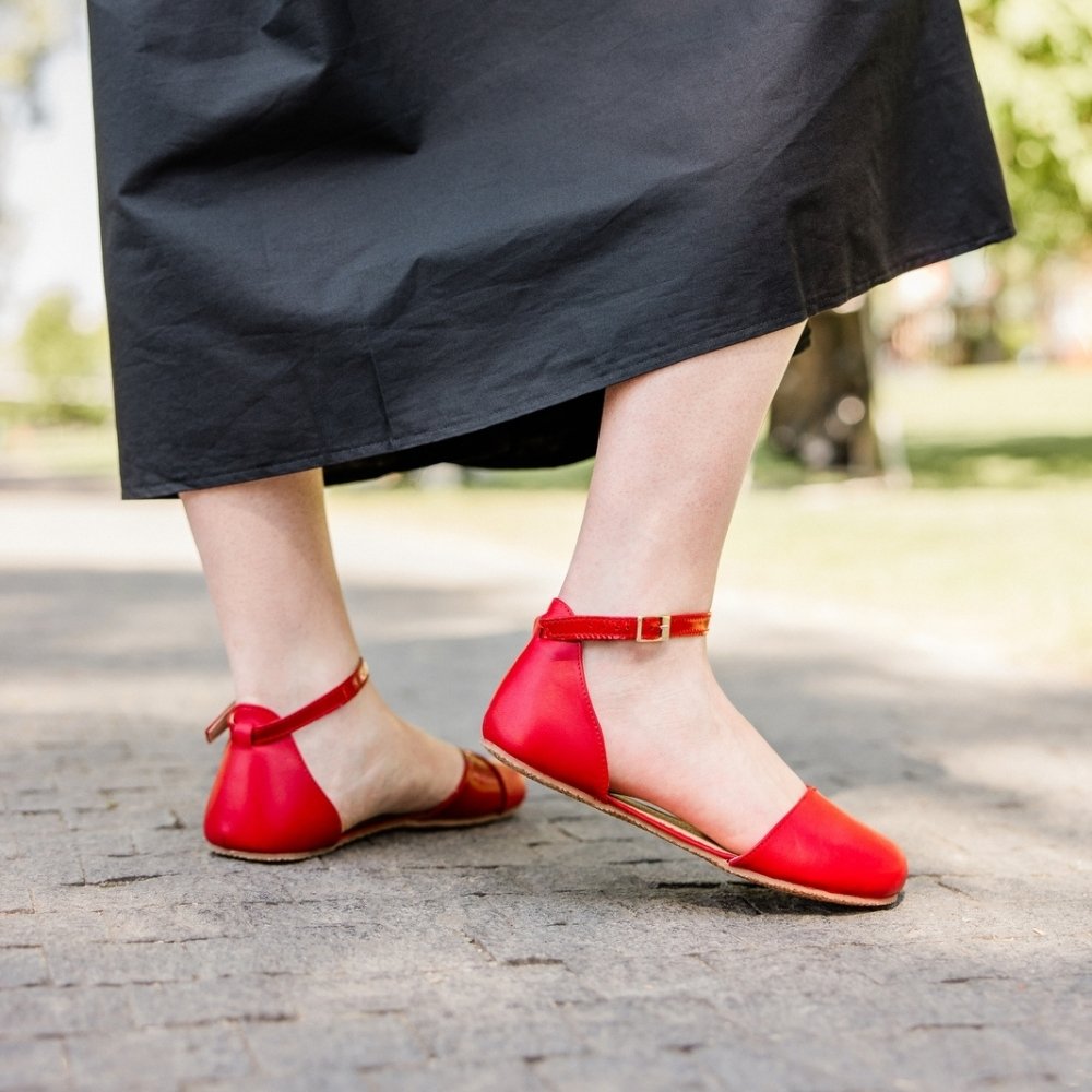Red leather Shapen Poppy D’orsay dressy flats with a tan footbed and tan rubber soles. The flats have a heel cup held on by a dainty, patent leather ankle strap. Both shoes are shown facing right on a light-skinned woman standing on a stone pathway wearing a long black dress with her right heel popped. #color_cherry