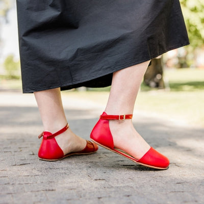 Red leather Shapen Poppy D’orsay dressy flats with a tan footbed and tan rubber soles. The flats have a heel cup held on by a dainty, patent leather ankle strap. Both shoes are shown facing right on a light-skinned woman standing on a stone pathway wearing a long black dress with her right heel popped. #color_cherry
