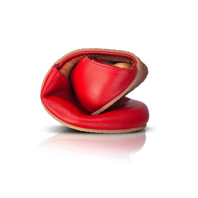 A photo of Shapen Poppy D’orsay dressy flats with a leather upper and rubber soles. The flats are a cherry red color with a heel cup and dainty ankle strap, the inside of the sole are a beige color. The right shoe is shown rolled in a ball against a white background. #color_red