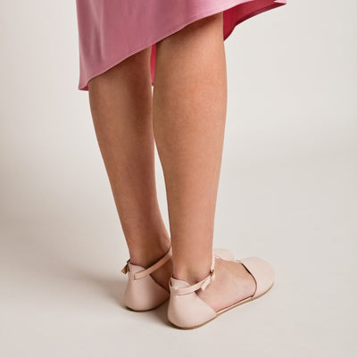A photo of Shapen Poppy D’orsay dressy flats with a leather upper and rubber soles. The flats are a rose beige color with a heel cup and dainty ankle strap, the inside of the sole are a beige color. Both flats are shown diagonally from the back right on a woman wearing a pink dress standing on a cream colored floor. #color_rose