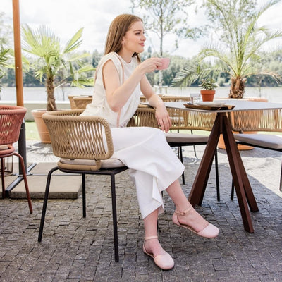 Rose leather Shapen Poppy D’orsay dressy flats with a tan footbed and tan rubber soles. The flats have a heel cup held on by a dainty, patent leather ankle strap. Both shoes are shown facing right on a light-skinned woman with short, light-brown hair. She is sitting in a cafe wearing a long white jumpsuit drinking coffee with her right leg crossed. #color_rose