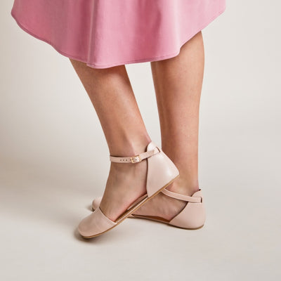 A photo of Shapen Poppy D’orsay dressy flats with a leather upper and rubber soles. The flats are a rose beige color with a heel cup and dainty ankle strap, the inside of the sole are a beige color. Both flats are shown from the left side on a woman wearing a pink dress with her left heel lifted standing on a cream colored floor. #color_rose