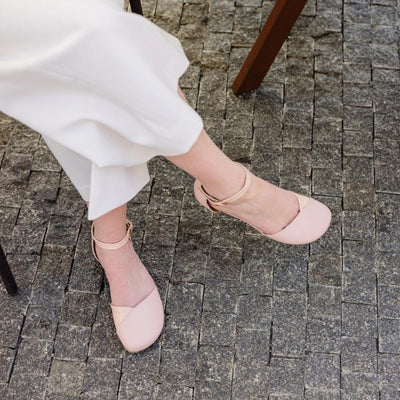 Rose leather Shapen Poppy D’orsay dressy flats with a tan footbed and tan rubber soles. The flats have a heel cup held on by a dainty, patent leather ankle strap. Both shoes are shown facing diagonally right on a light-skinned woman sitting in a cafe wearing a long white jumpsuit drinking coffee with her right leg crossed. #color_rose