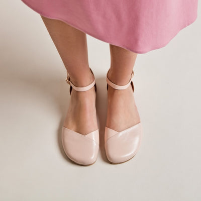A photo of Shapen Poppy D’orsay dressy flats with a leather upper and rubber soles. The flats are a rose beige color with a heel cup and dainty ankle strap, the inside of the sole are a beige color. Both flats are shown from the top down on a woman wearing a pink dress standing on a cream colored floor. #color_rose