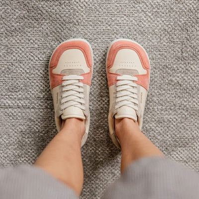 Peach Shapen ReWind leather sneakers. Shoes are a classic chunky sneaker design with light pink leather and peach suede color blocks surrounding the toe guard and heels. Light grey suede color blocks are on the perimeter of the laces and the top of the heel. Both shoes are shown from above on a person wearing loose grey shorts standing on a carpet. #color_peach