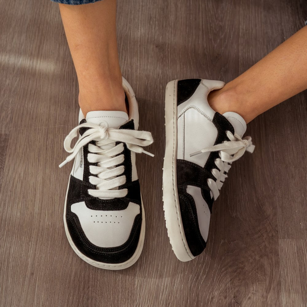 A photo of Shapen ReWind leather sneakers in black & white. Shoes are a classic chunky sneaker design with black suede color blocks surrounding the toe guard, laces, and heels. Both shoes are shown here from the top down with the left shoe tilted to the left. Shoes are on a womans feet wearing medium wash loose blue jeans sitting on a grey wood floor. #color_black-white