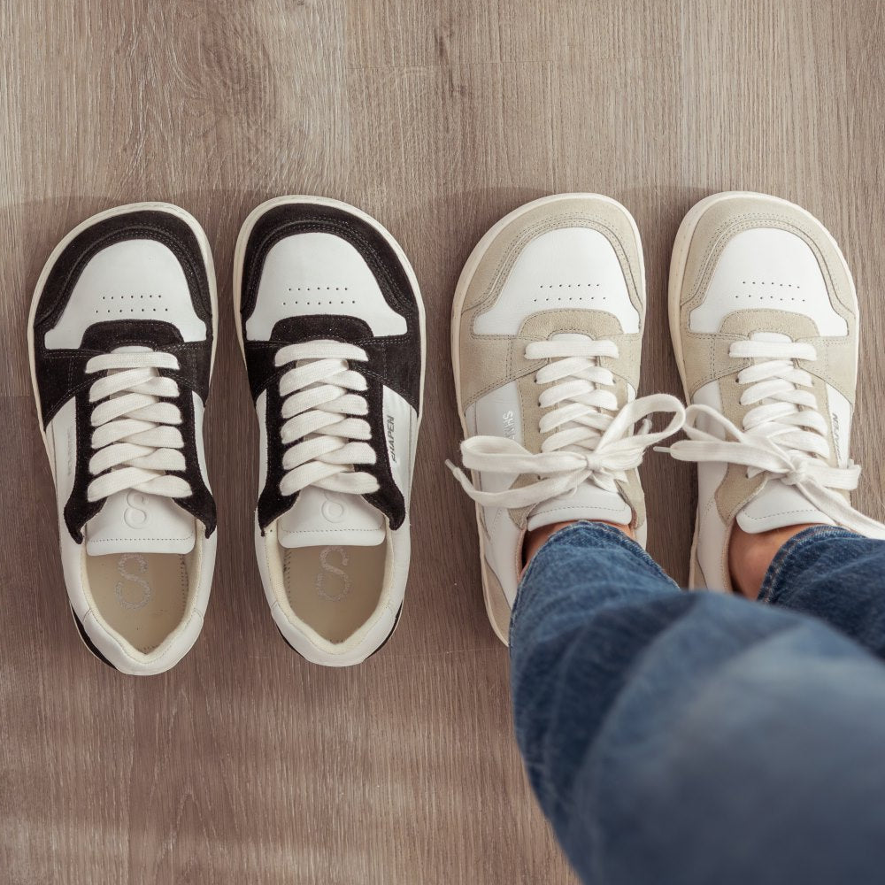 A photo of Shapen ReWind leather sneakers in white. Shoes are a classic chunky sneaker design with beige suede color blocks surrounding the toe guard, laces, and heels. Both shoes are shown here from the top down. Shoes are on a womans feet wearing medium wash loose blue jeans standeing on a grey wood floor. ReWind in White & Black are lined up next to the left of the White shoes. #color_white