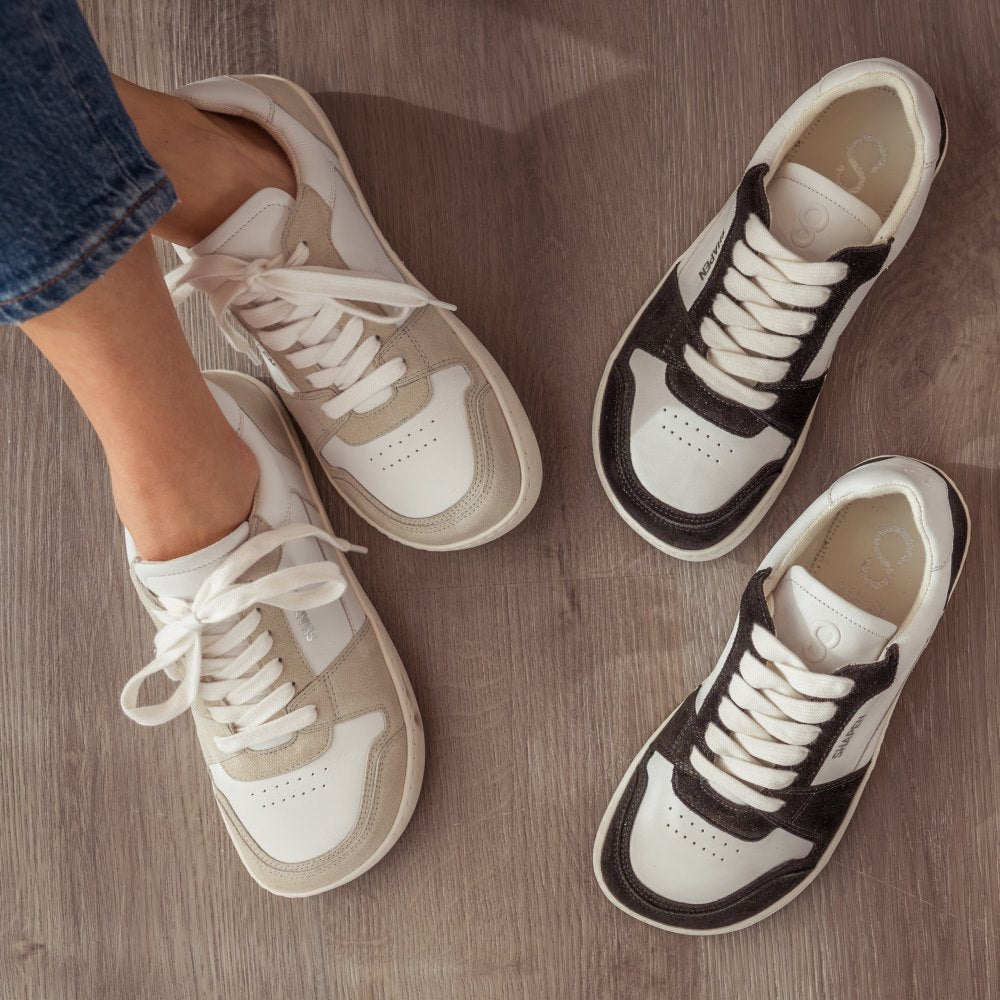 A photo of Shapen ReWind leather sneakers in white. Shoes are a classic chunky sneaker design with beige suede color blocks surrounding the toe guard, laces, and heels. Both shoes are shown here from the top down with the left shoe crossed over the right. Shoes are on a womans feet wearing medium wash loose blue jeans sitting on a grey wood floor. ReWind in White & Black are staggered to the right of the White shoes. #color_black-white
