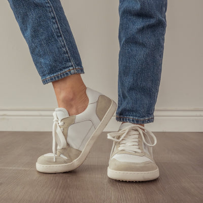 A photo of Shapen ReWind leather sneakers in white. Shoes are a classic chunky sneaker design with beige suede color blocks surrounding the toe guard, laces, and heels. Left shoe is shown here turned to the left while right shoe is facing the camera. Shoes are on a womans feet wearing medium wash loose blue jeans. #color_white