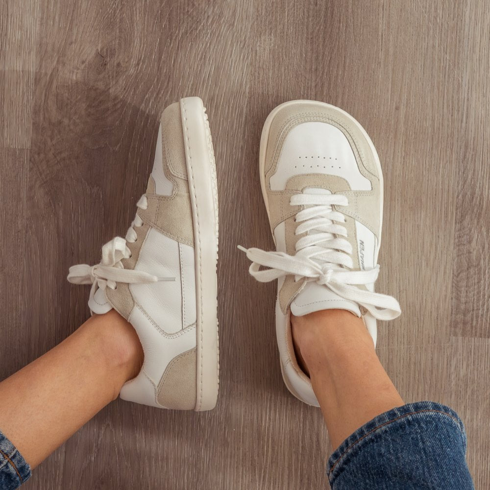 A photo of Shapen ReWind leather sneakers in white. Shoes are a classic chunky sneaker design with beige suede color blocks surrounding the toe guard, laces, and heels. Both shoes are shown here from the top down with the left shoe tilted to the left. Shoes are on a womans feet wearing medium wash loose blue jeans sitting on a grey wood floor. #color_white