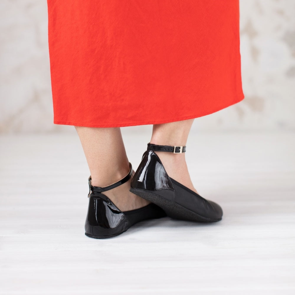 A photo of Shapen Tulip flats in black made of leather and rubber soles. Shoes have a gentle elastic around the perimeter of the opening, a patent leather heel cup, and an optional patent leather ankle strap. Both shoes are shown here from the back worn by a woman wearing a bright red, mid-length, tank top dress with side slits standing on a white wooden floor. #color_black