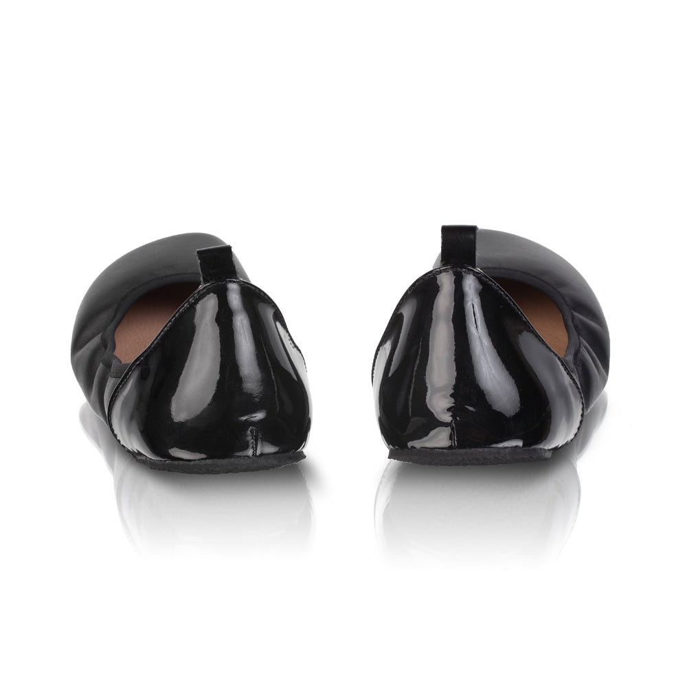 A photo of Shapen Tulip flats in black made of leather and rubber soles. Shoes have a gentle elastic around the perimeter of the opening, a patent leather heel cup, and an optional patent leather ankle strap. Both shoes are shown here without the ankle strap from the back against a white background. #color_black