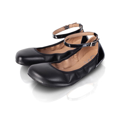 A photo of Shapen Tulip flats in black made of leather and rubber soles. Shoes have a gentle elastic around the perimeter of the opening, a patent leather heel cup, and an optional patent leather ankle strap. Both shoes are shown diagonally here from the front left against a white background. #color_black