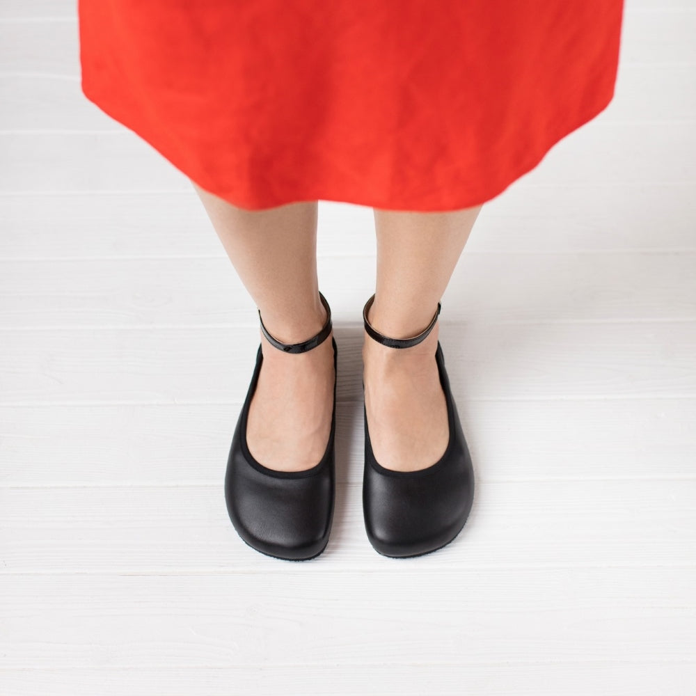 A photo of Shapen Tulip flats in black made of leather and rubber soles. Shoes have a gentle elastic around the perimeter of the opening, a patent leather heel cup, and an optional patent leather ankle strap. Both shoes are shown here from the top down worn by a woman wearing a bright red, mid-length, tank top dress with side slits standing on a white wooden floor. #color_black