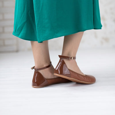 A photo of Shapen Tulip flats in cognac made of leather and rubber soles. Shoes have a gentle elastic around the perimeter of the opening, a patent leather heel cup, and an optional patent leather ankle strap. Both shoes are shown here from the back left side worn by a woman wearing a muted emerald green, mid-length, long sleeve dress with waist slits standing on a white wooden floor with the left right bent slightly. #color_cognac