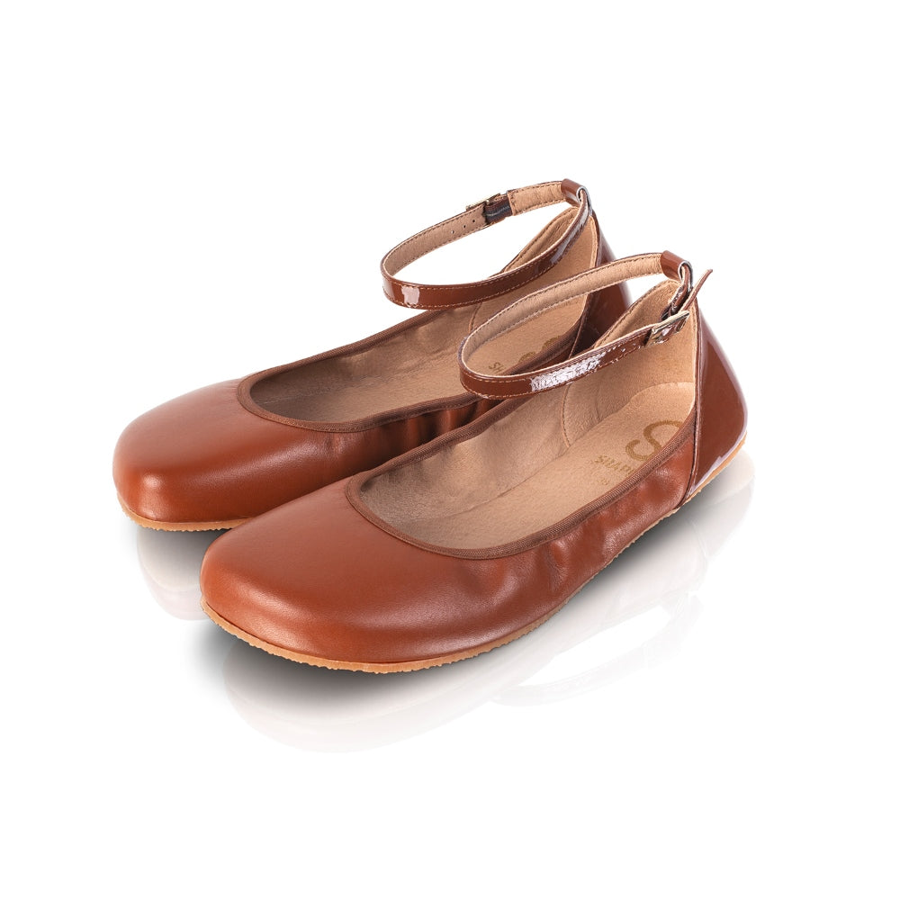 A photo of Shapen Tulip flats in cognac made of leather and rubber soles. Shoes have a gentle elastic around the perimeter of the opening, a patent leather heel cup, and an optional patent leather ankle strap. Both shoes are shown diagonally here from the front left against a white background. #color_cognac