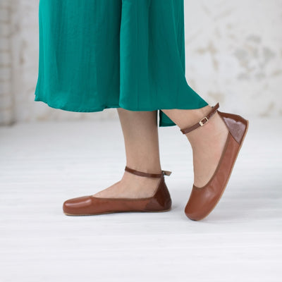 A photo of Shapen Tulip flats in cognac made of leather and rubber soles. Shoes have a gentle elastic around the perimeter of the opening, a patent leather heel cup, and an optional patent leather ankle strap. Both shoes are shown here from the left side worn by a woman wearing a muted emerald green, mid-length, long sleeve dress with waist slits standing on a white wooden floor with the left leg bent and toe box resting on the floor. #color_cognac