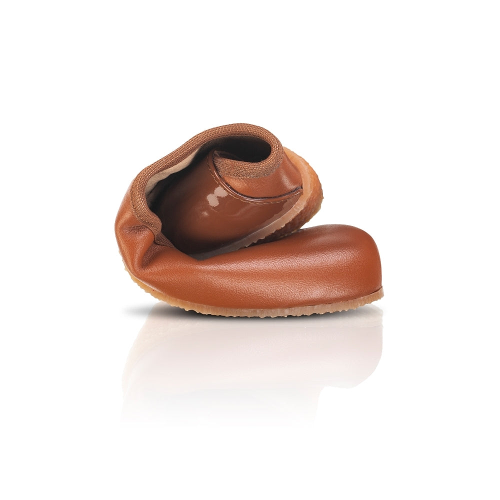A photo of Shapen Tulip flats in cognac made of leather and rubber soles. Shoes have a gentle elastic around the perimeter of the opening, a patent leather heel cup, and an optional patent leather ankle strap. One shoe is shown rolled here against a white background. #color_cognac