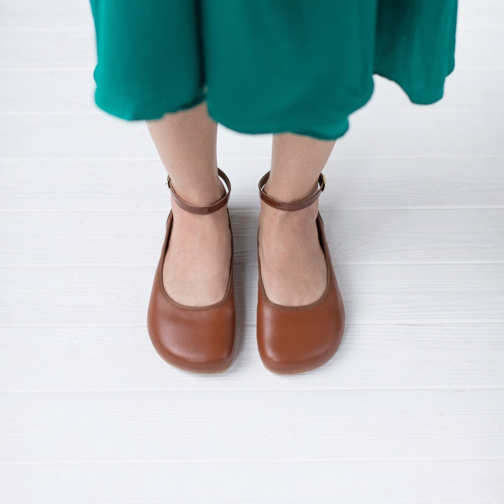 A photo of Shapen Tulip flats in cognac made of leather and rubber soles. Shoes have a gentle elastic around the perimeter of the opening, a patent leather heel cup, and an optional patent leather ankle strap. Both shoes are shown here from the top down worn by a woman wearing a muted emerald green, mid-length, long sleeve dress with waist slits standing on a white wooden floor. #color_cognac