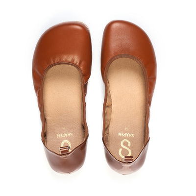 A photo of Shapen Tulip flats in cognac made of leather and rubber soles. Shoes have a gentle elastic around the perimeter of the opening, a patent leather heel cup, and an optional patent leather ankle strap. Both wide width shoes are shown here without the ankle strap from the top down against a white background. #color_cognac