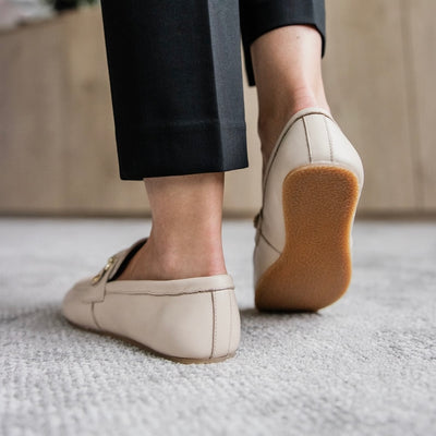 Beige leather Shapen Versa loafers. Shoes are a classic loafer design with a leather decoration capped by gold metal rings across the foot and tan rubber soles glued on. Both shoes are from behind on a light skinned woman wearing cropped black slacks standing on a white carpet with her right foot tilted to show the sole of the shoe. #color_beige