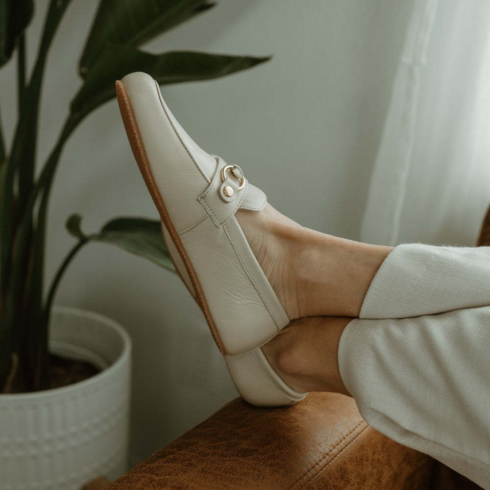 Beige leather Shapen Versa loafers. Shoes are a classic loafer design with a leather decoration capped by gold metal rings across the foot and tan rubber soles glued on. Both shoes are shown on a light skinned woman wearing wide leg ivory slacks sitting on a cognac leather couch with her feet propped on the left arm rest and a plant in the background. #color_beige