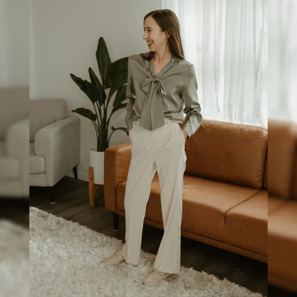 Beige leather Shapen Versa loafers. Shoes are a classic loafer design with a leather decoration capped by gold metal rings across the foot and tan rubber soles glued on. Both shoes are shown facing diagonally left on a light skinned woman with long brown straight hair. She is wearing wide leg ivory slacks with a sage satin long sleeve shirt with a tie collar tied into a bow standing on a plush ivory carpet. A cognac leather couch, tall plant, and tan chair are in the background. #color_beige