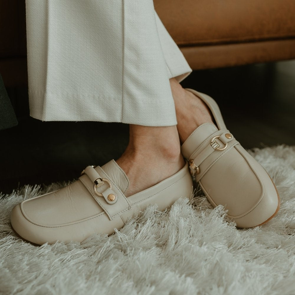 Beige leather Shapen Versa loafers. Shoes are a classic loafer design with a leather decoration capped by gold metal rings across the foot and tan rubber soles glued on. Both shoes are shown facing diagonally left on a light skinned woman wearing wide leg ivory slacks sitting on a cognac leather couch with a plush ivory carpet below. #color_beige