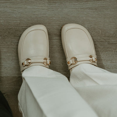 Beige leather Shapen Versa loafers. Shoes are a classic loafer design with a leather decoration capped by gold metal rings across the foot and tan rubber soles glued on. Both shoes are shown from above against a grey wooden floor. #color_beige