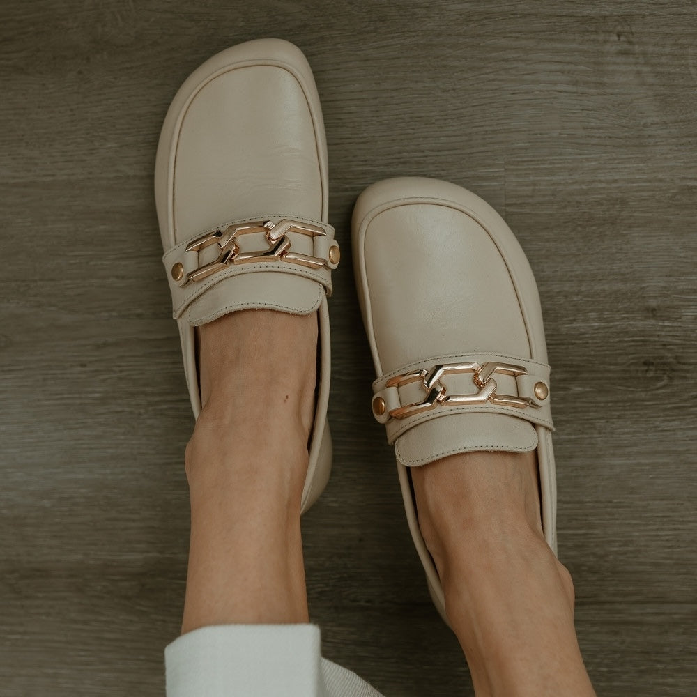 A hexagonal 3 link gold chain accessory for the Shapen Versa Loafer. Chain is shown from above on the beige Versa loafer on a person wearing loose white slacks sitting on a grey wooden floor.  #color_gold-chain