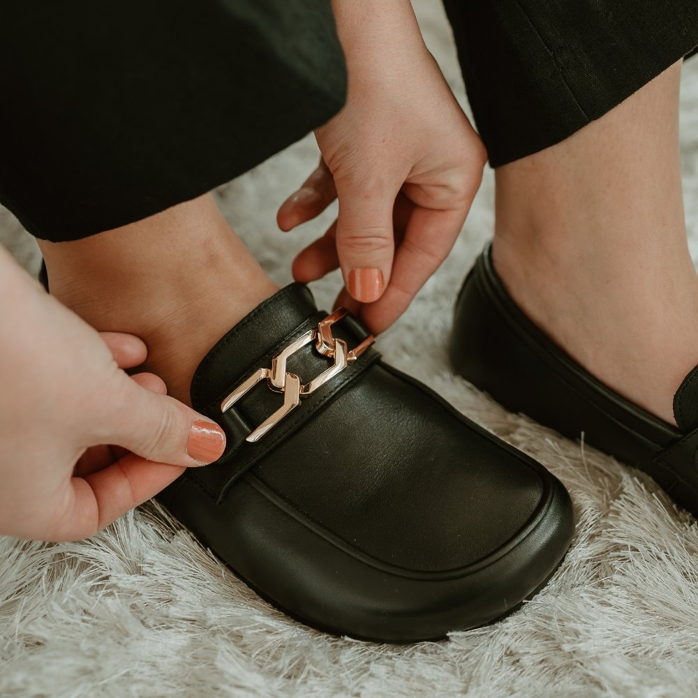 A hexagonal 3 link gold chain accessory for the Shapen Versa Loafer. Chain is shown facing diagonally right on the black Versa loafer on a person wearing loose black slacks standing on a fluffy white carpet. #color_gold-chain