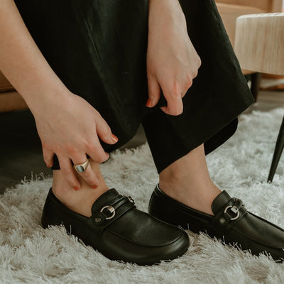 Black leather Shapen Versa loafers. Shoes are a classic loafer design with a leather decoration capped by silver metal rings across the foot and black rubber soles glued on.Both shoes are shown facing diagonally right on a light skinned woman wearing wide leg black slacks holding her pant legs on a plush white carpet. #color_black
