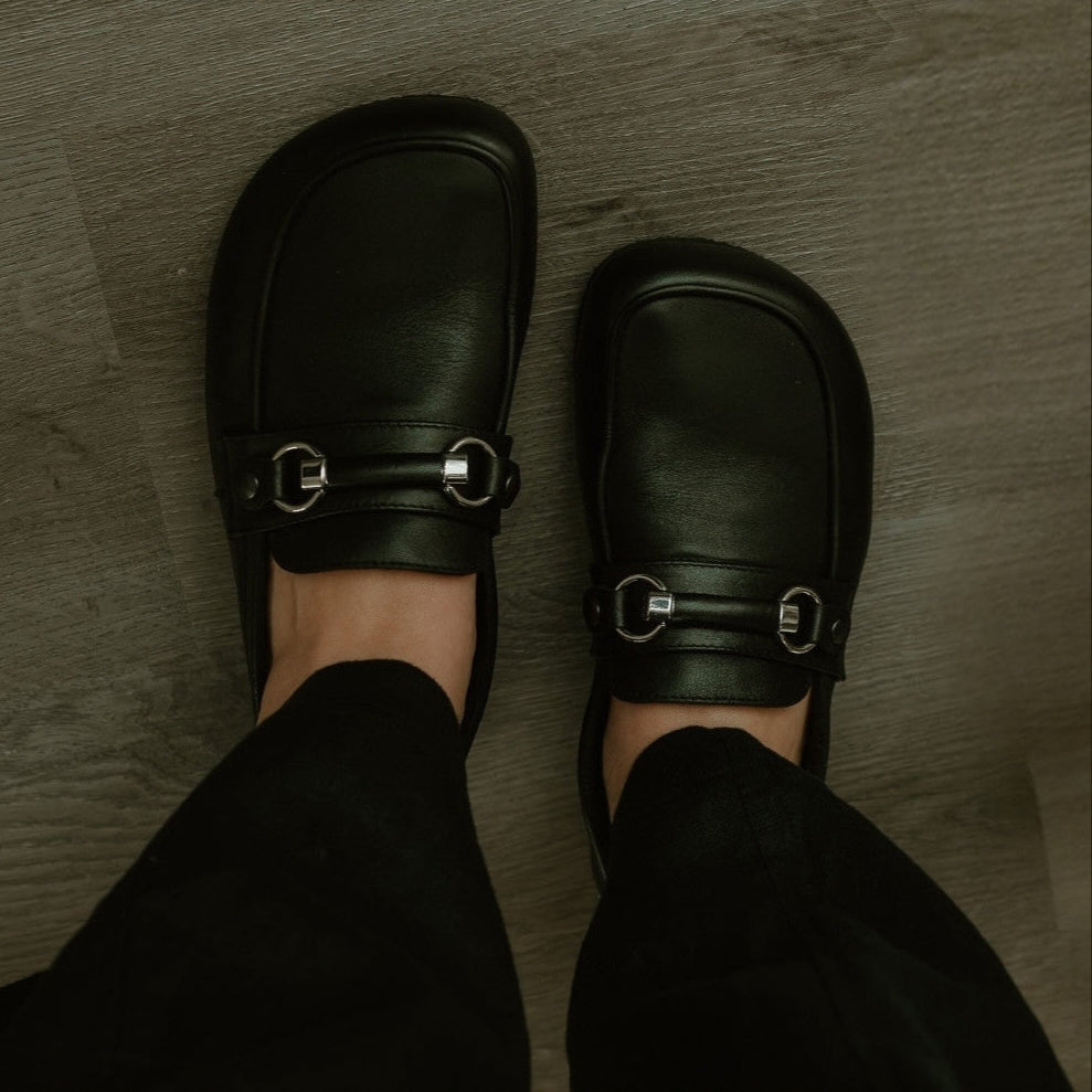 Black leather Shapen Versa loafers. Shoes are a classic loafer design with a leather decoration capped by silver metal rings across the foot and black rubber soles glued on. Both shoes are shown from above on a light skinned woman wearing wide leg black slacks standing on a grey wooden floor. #color_black