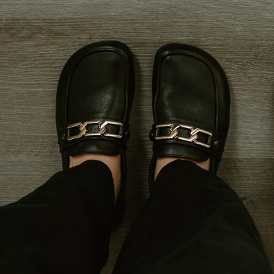 A hexagonal 3 link gold chain accessory for the Shapen Versa Loafer. Chain is shown from above on the black Versa loafer on a person wearing loose black slacks standing on a grey wooden floor.  #color_gold-chain
