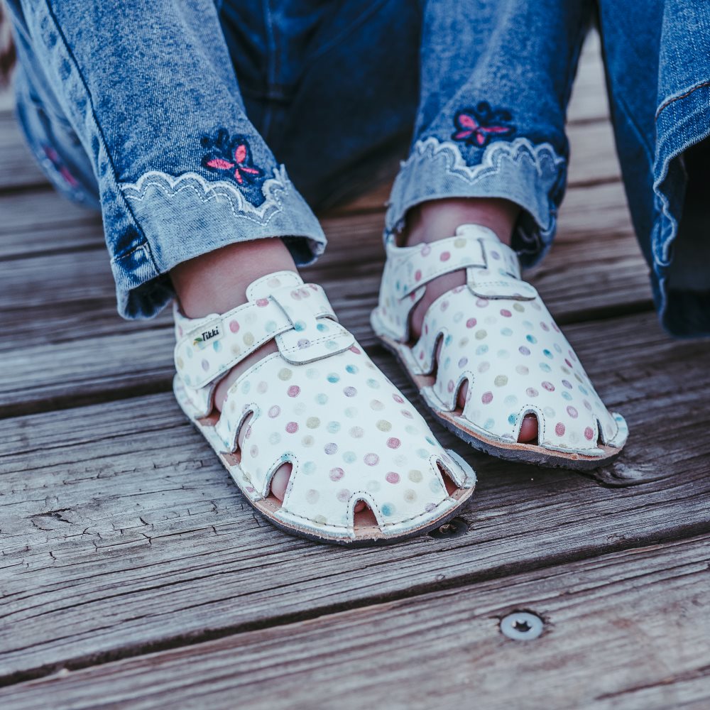 A photo of Tikki Aranya kids sandals in Confetti. Sandals are surrounded in leather with 4 slits on each side and a velcro closure across the foot. Footbed is brown and soles are black. Both shoes are shown here facing diagonally to the right on a little girl wearing skinny jeans with lace detail and a pink embroidered butterfly at the cuff sitting on a wooden dock. #color_confetti