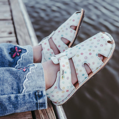 A photo of Tikki Aranya kids sandals in Confetti. Sandals are surrounded in leather with 4 slits on each side and a velcro closure across the foot. Footbed is brown and soles are black. Both shoes are shown here from the right on a little girl wearing skinny jeans with lace detail and a pink embroidered butterfly at the cuff sitting on a wooden dock with her feet hanging parallel above the water. #color_confetti