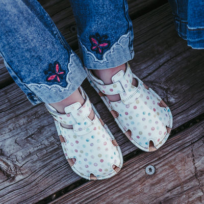 A photo of Tikki Aranya kids sandals in Confetti. Sandals are surrounded in leather with 4 slits on each side and a velcro closure across the foot. Footbed is brown and soles are black. Both shoes are shown here from above facing diagonally to the right on a little girl wearing skinny jeans with lace detail and a pink embroidered butterfly at the cuff sitting on a wooden dock. #color_confetti