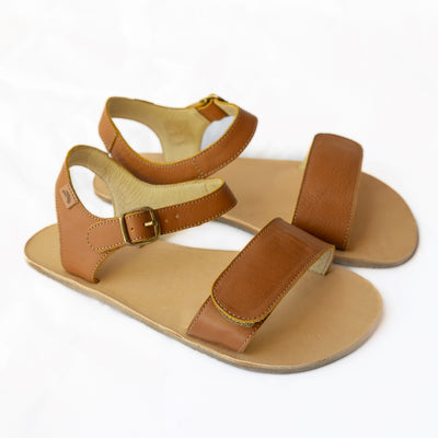 Tikki Vibe brown leather sandals with a tan footbed and soles. A thick velcro strap secures the foot at the toes with a slimmer ankle buckle-closure strap in the back. Both shoes are shown here facing diagonally right against a white background. #color_cream-leather