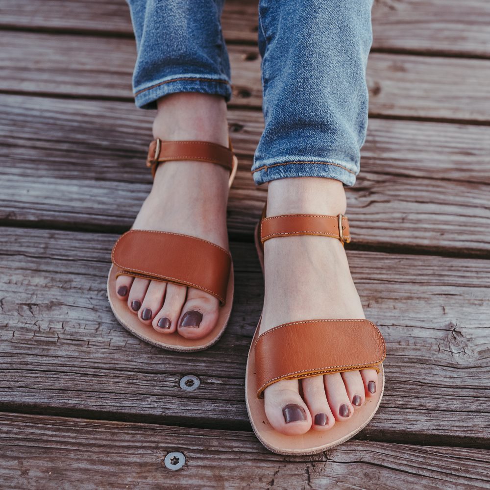 Tikki Vibe brown leather sandals with a tan footbed and cream vibram soles. A thick velcro strap secures the foot at the toes with a slimmer ankle buckle-closure strap in the back. Both shoes are shown here from the front on a young lady wearing flare jenas sitting on a wooden dock. #color_cream-leather