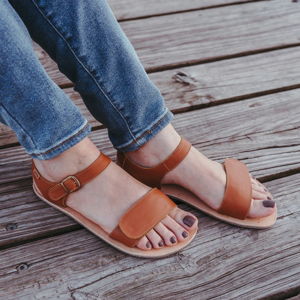 Tikki Vibe brown leather sandals with a tan footbed and cream vibram soles. A thick velcro strap secures the foot at the toes with a slimmer ankle buckle-closure strap in the back. Both shoes are shown here facing diagonally right on a young lady wearing flare jenas sitting on a wooden dock. #color_cream-leather