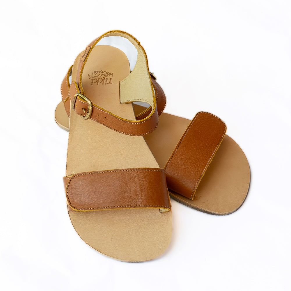 Tikki Vibe brown leather sandals with a tan footbed and soles. A thick velcro strap secures the foot at the toes with a slimmer ankle buckle-closure strap in the back. Both shoes are shown here from the front with the right shoe propped up on the left against a white background. #color_cream-leather