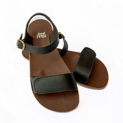 Tikki Vibe black vegan sandals with tan soles and a brown footbed. A thick velcro strap secures the foot at the toes with a slimmer ankle buckle-closure strap in the back. Right shoe is shown here from the front resting on the left shoe facing left against a white background. #color_onyx-vegan