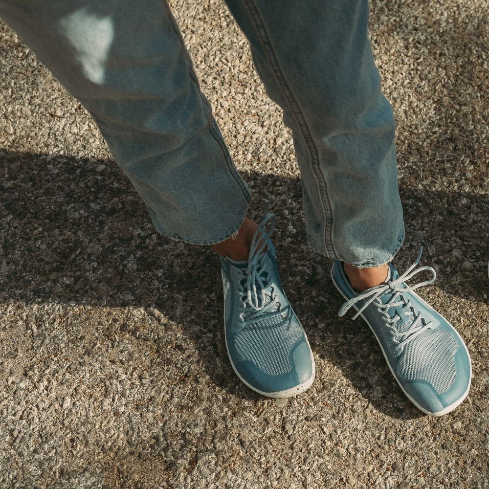 A photo of Vivobarefoot Primus lite sneakers made from recycled plastic and a rubber sole. The athletic sneakers are a blue haze color with white soles spotted with color with the vivobarefoot logo on the side. Both shoes are shown from above facing diagonally right on a tan woman wearing light-wash, loose-fitting jeans standing on pavement. #color_blue-haze