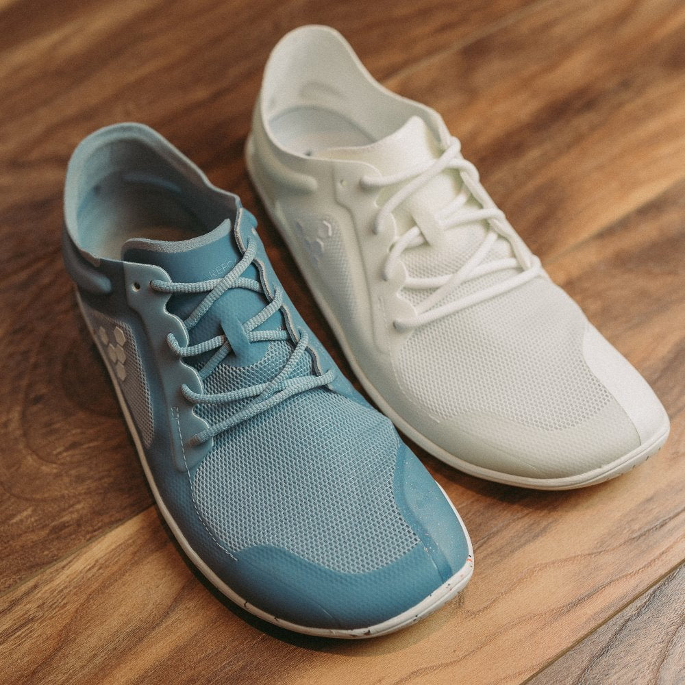 A photo of Vivobarefoot Primus lite sneakers made from recycled plastic and a rubber sole. The athletic sneakers are a blue haze color with white soles spotted with color and the vivobarefoot logo on the side. Right shoe is shown here next to a right, bright white primus lite. Both shoes are facing diagonally to the right on a wooden floor. #color_blue-haze