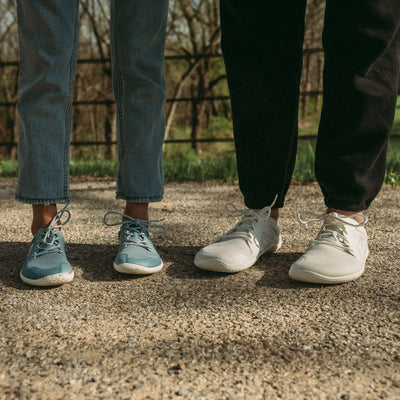 A photo of Vivobarefoot Primus lite sneakers made from recycled plastic and a rubber sole. The athletic sneakers are a blue haze color with white soles spotted with color with the vivobarefoot logo on the side. Both shoes are shown from the front on a tan woman wearing light-wash, loose-fitting jeans standing on pavement next to a person wearing black joggers and the bright white version of the sneaker. #color_blue-haze